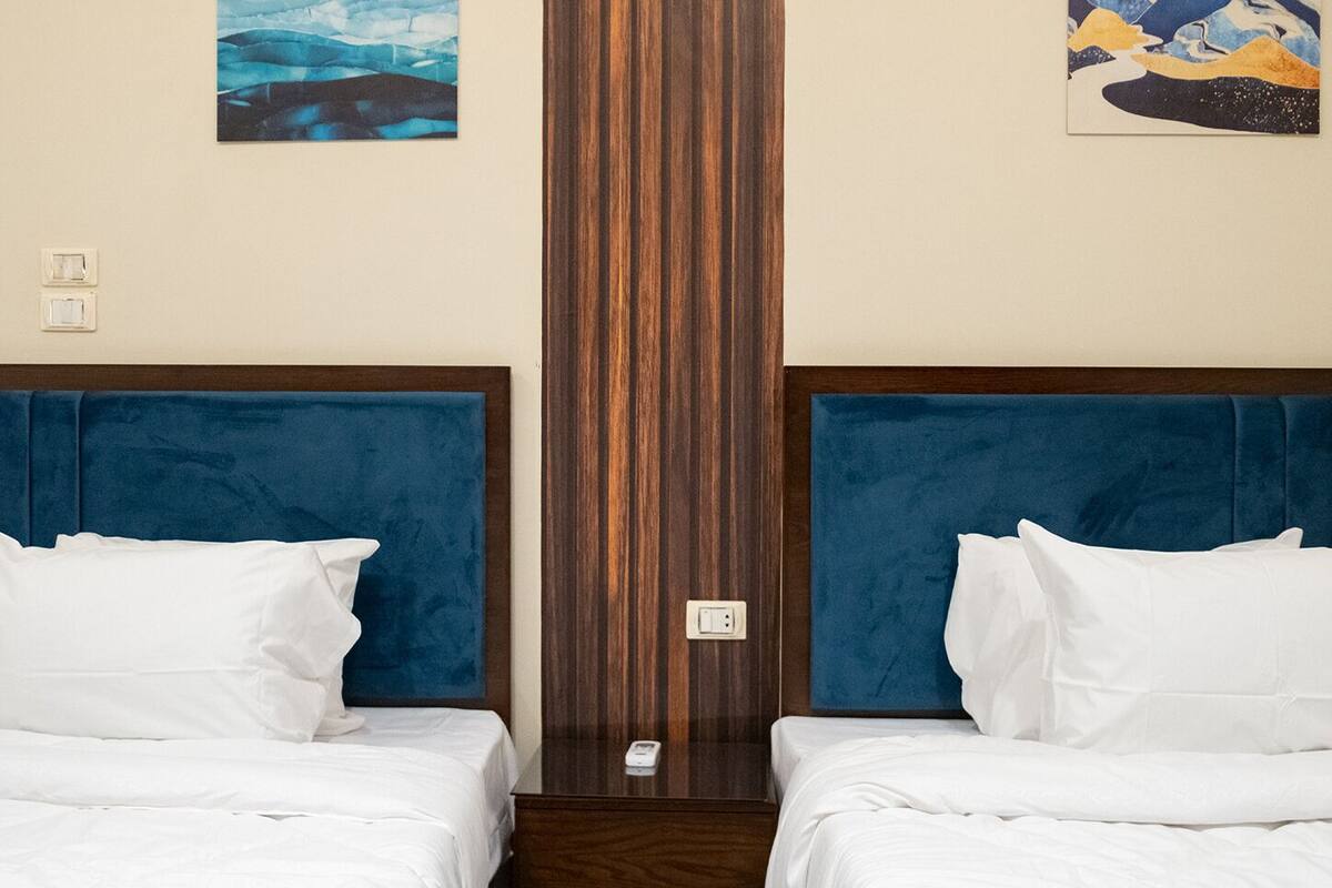 A hotel room with two beds, each with a blue headboard, white bedding, and a wooden panel in the center.