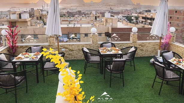 A rooftop dining area with tables and chairs, a view of the pyramids, and the Alba logo. - Alba pyramids view inn (Giza)