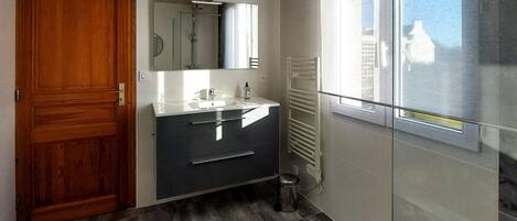 A bathroom with a wooden door, a sink with a mirror, a towel rack, and a window with a view of buildings.