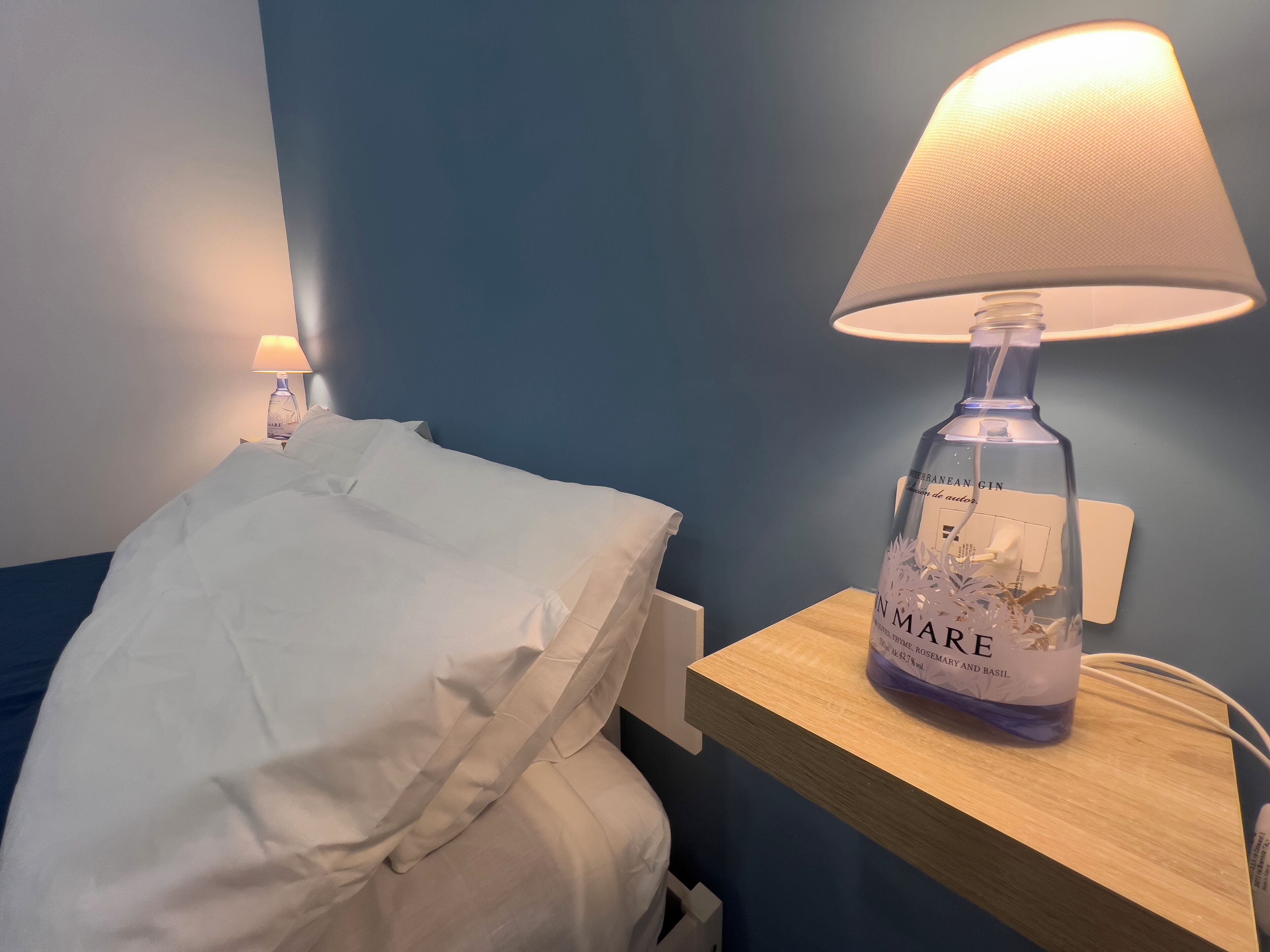A bed with white bedding, a bedside lamp, and a bottle of In Marque herbal gin on a wooden nightstand.
