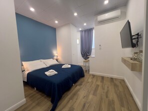 A hotel room with a bed, a nightstand, a desk, a chair, an air conditioning unit, and a mounted TV. - Aura apartments (Naples)