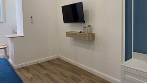 Un dormitorio con una televisión montada en la pared, una pequeña estantería con objetos decorativos y suelo de madera.