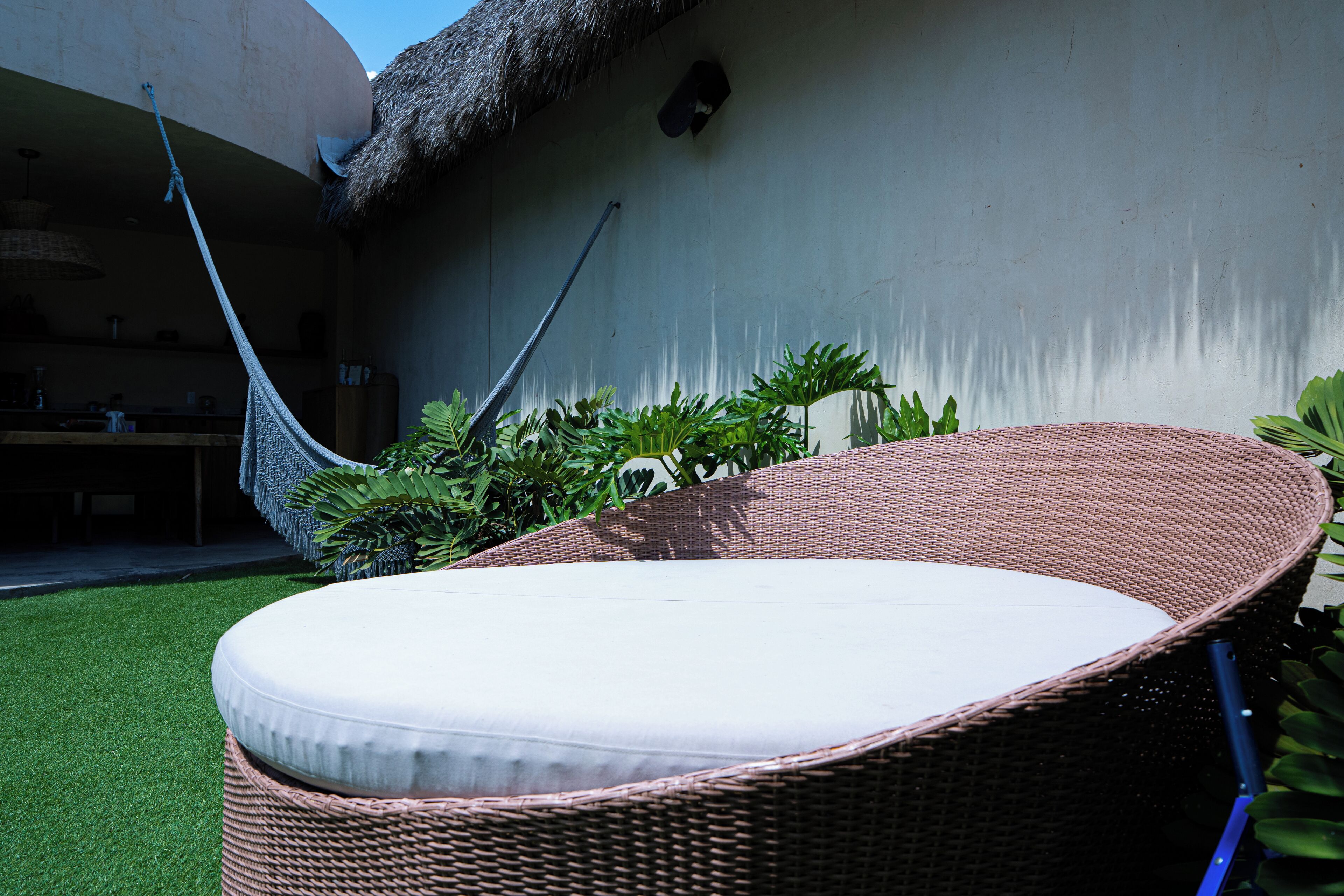 A wicker lounge chair with a white cushion in a garden area with a thatched roof structure and green foliage.