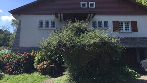 A two-story building with a red roof and white walls, featuring a small porch and a garden with flowering plants. - Large, comfortable house in Bagneres  (Bagnères-de-Bigorre)