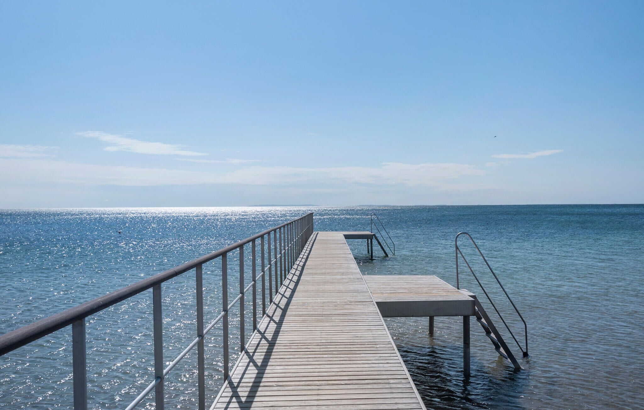 Une jetée en bois s’étendant dans des eaux calmes, avec une rambarde métallique d’un côté et un escalier métallique de l’autre.