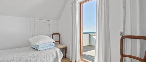A small, cozy bedroom with a single bed, a chair, and a view of the sea through a sliding glass door.
