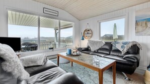 Un salon confortable avec un canapé en cuir, une table basse et un téléviseur. De grandes fenêtres offrent une vue sur l’extérieur.