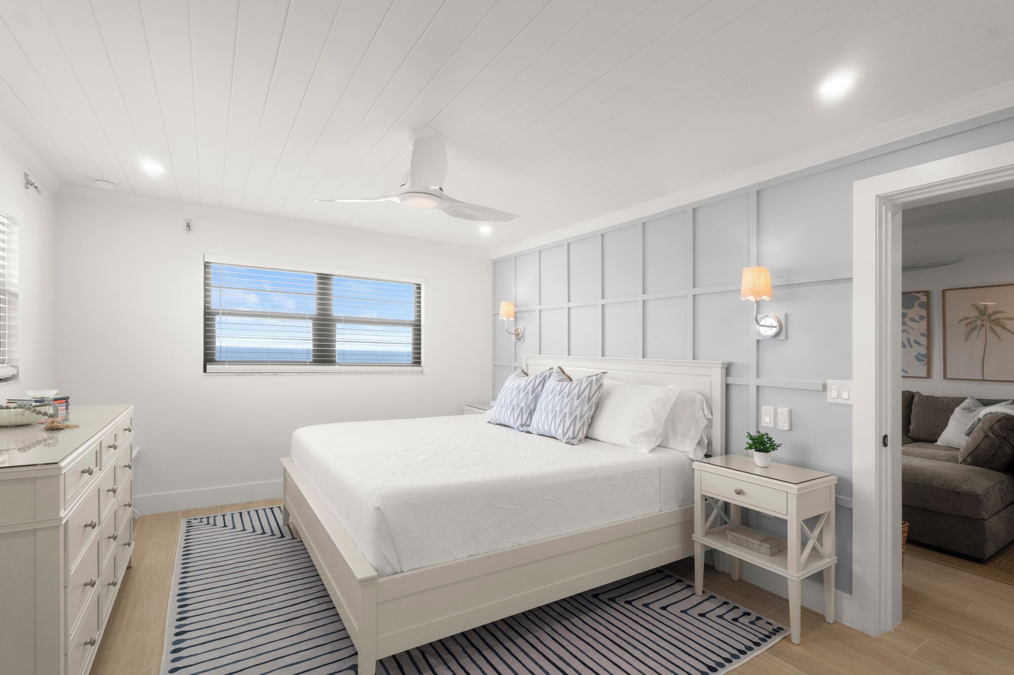 A bedroom with a bed, bedside tables, a nightstand, a window with blinds, a ceiling fan, and a view of the sea.