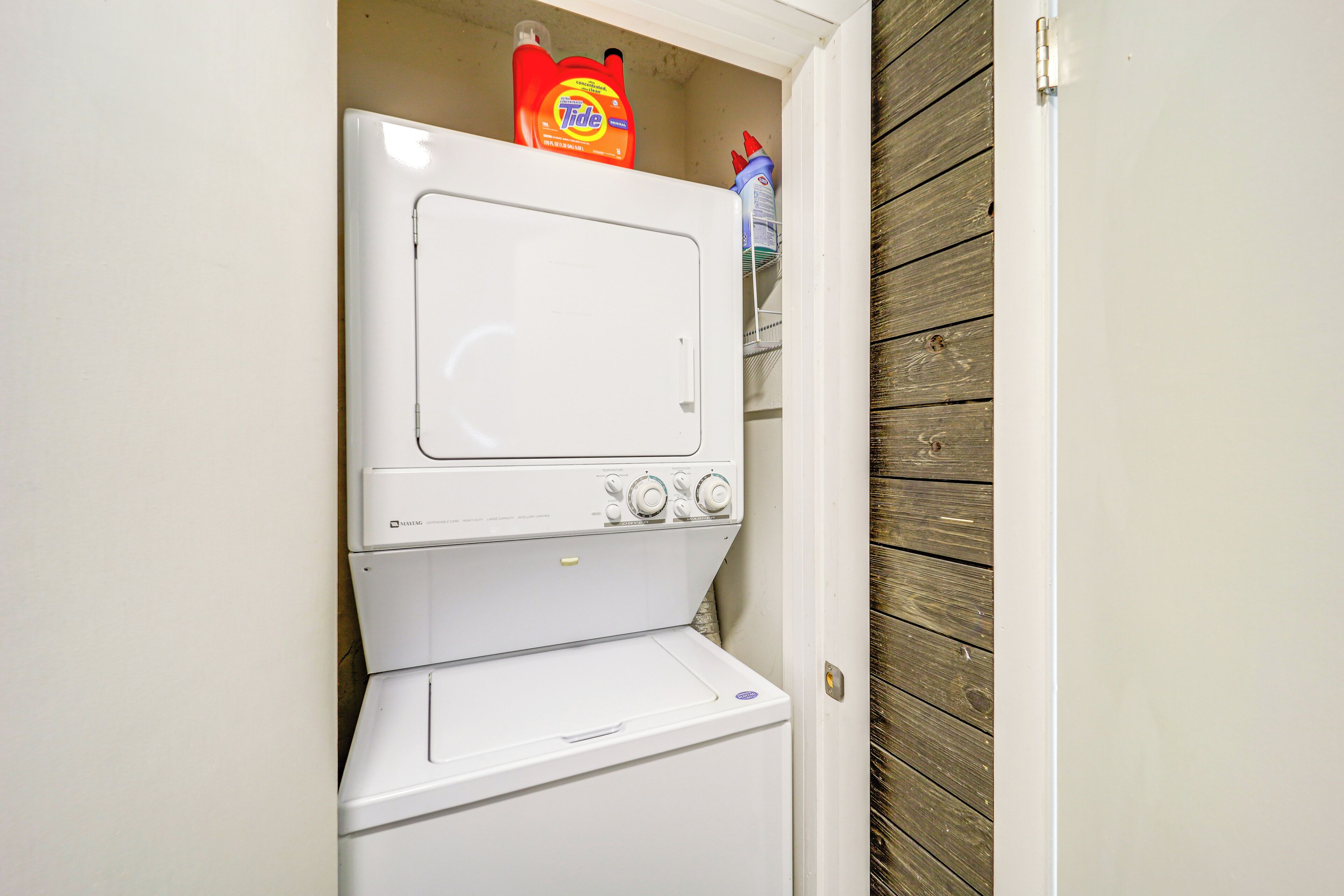A compact laundry room with a front-loading washer and dryer, a shelf with Tide detergent, and a wooden wall.