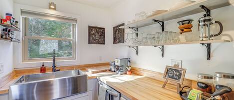 A modern kitchen with a stainless steel sink, wooden countertop, and shelves holding glassware and kitchen items.
