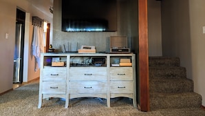 Una habitación con escalera de madera, un televisor de pantalla plana montado en la pared, un mueble de madera con cajones y una zona de cocina con refrigerador y microondas.