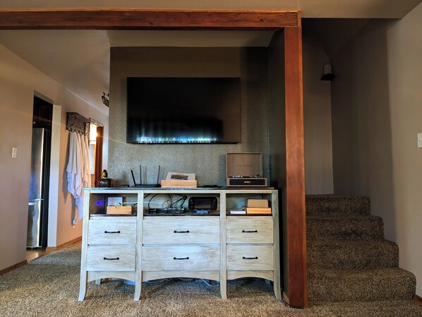 Una habitación con escalera de madera, un televisor de pantalla plana montado en la pared, un mueble de madera con cajones y una zona de cocina con refrigerador y microondas.