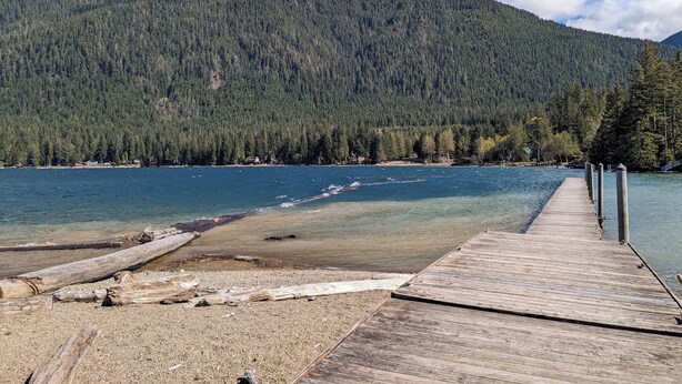 Muellé de madera que se adentra en un lago tranquilo de aguas cristalinas, rodeado de un bosque denso y montañas al fondo.