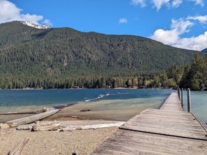 A wooden dock extending into a calm lake with clear water, surrounded by dense forest and mountains in the background.