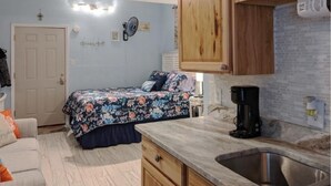 A compact bedroom with a bed, a bedside table, and a nightstand. There is a kitchenette area with a sink, a stove, and a coffee maker.