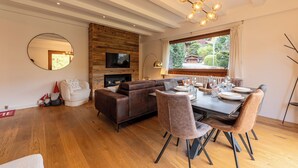 A modern living room with a wooden wall, a round mirror, a brown sofa, a dining table with chairs, and a TV mounted on the wall.