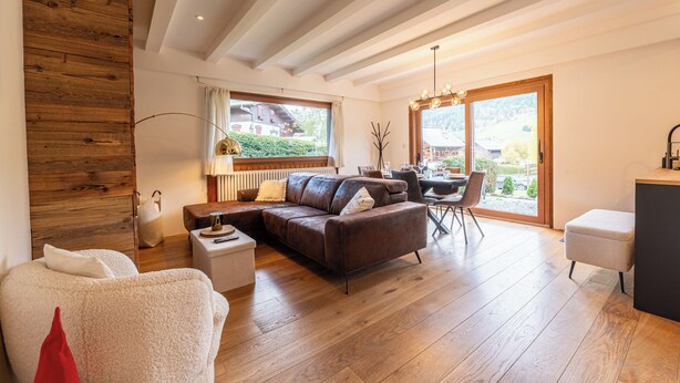 A modern living room with a wooden ceiling, a brown leather sofa, a white armchair, and a dining area with a table and chairs.