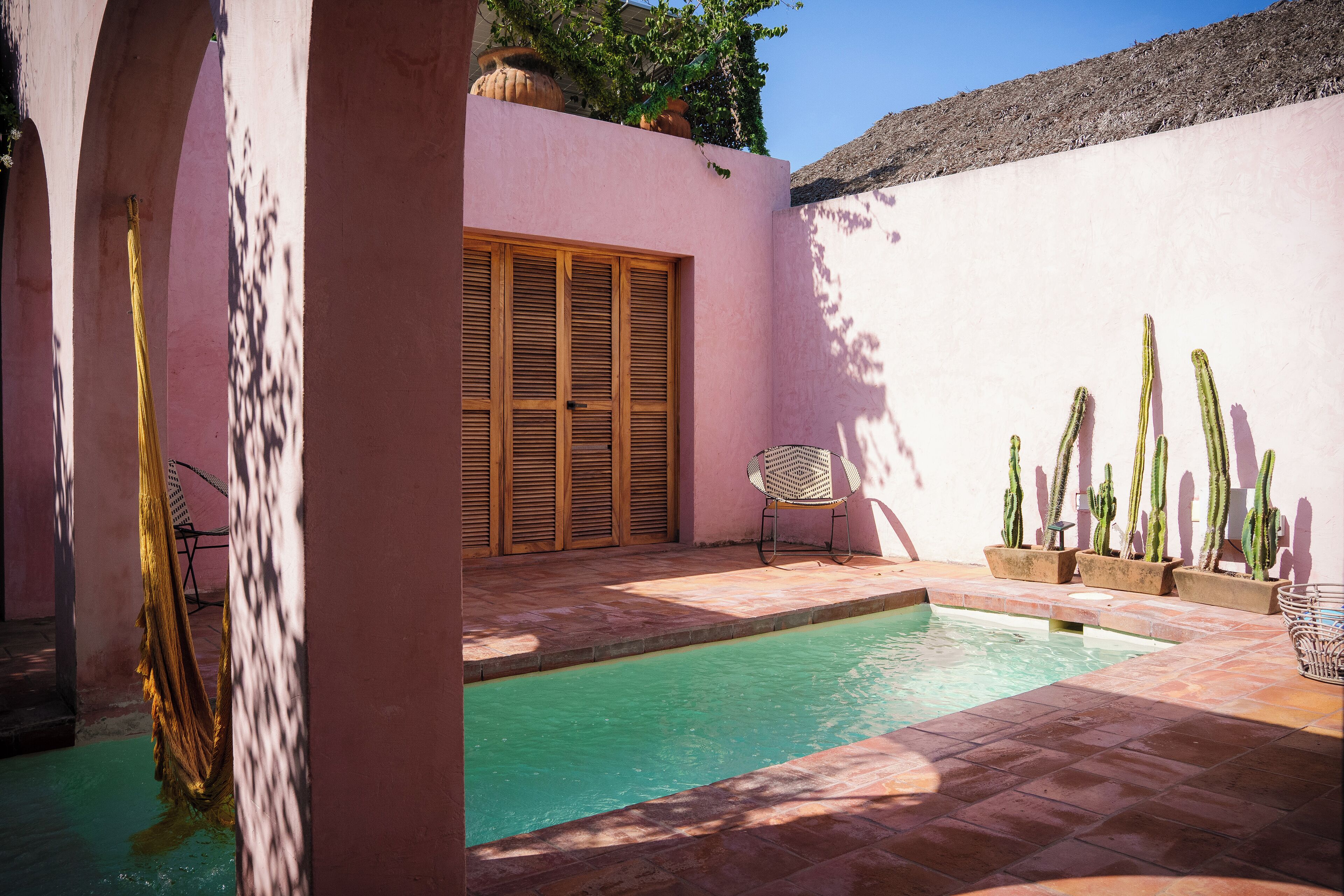 Zona de piscina con pared rosa, contraventanas de madera, una silla y cactus.