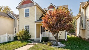 A two-story residential building with a front porch, a small tree with autumn leaves, and a white picket fence. - Charming Retreat with Private Patio - Cozy and Convenient Escape (Indianapolis)