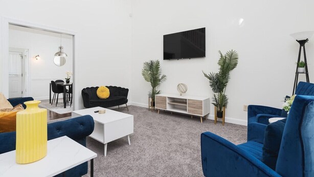 A modern living room with a white sofa, a blue sofa, a white coffee table, and a black sofa with a yellow pillow. There is a TV mounted on the wall, a small white cabinet, and two potted plants. - Charming Retreat with Private Patio - Cozy and Convenient Escape (Indianapolis)