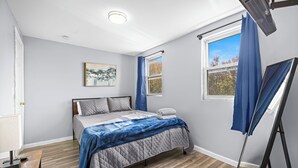Un dormitorio con cama, un cuadro, una ventana con cortinas y un ventilador de pie.