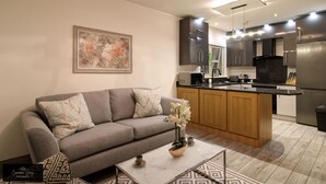 Una sala de estar moderna con un sofá gris, una mesa de centro con una planta, y una cocina con refrigerador, microondas y armarios de madera.