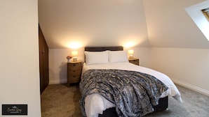 Un dormitorio con techo inclinado, una cama con manta estampada, dos lámparas de noche y una mesita de noche con un florero.
