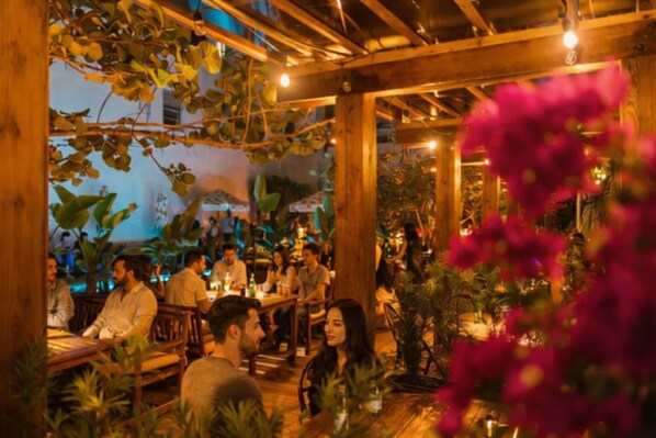 An outdoor dining area with wooden beams, tables, and chairs, surrounded by greenery and plants.