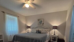A bedroom with a bed, two bedside tables with lamps, a ceiling fan, a window with blinds, and a small rug.