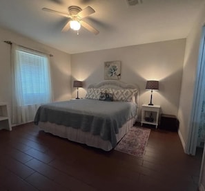 A bedroom with a bed, two bedside tables with lamps, a ceiling fan, a window with blinds, and a small rug.