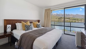 Ein Hotelzimmer mit einem großen Bett, einem Nachttisch, einer Kommode und Blick auf einen See und Berge.