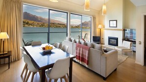Ein modernes Wohnzimmer mit großem Fenster mit Blick auf einen See und Berge, einem Essbereich mit Tisch und Stühlen und einer gemütlichen Sitzecke mit Sofa und Kamin.
