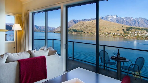 Ein Hotelzimmer mit großem Fenster und Blick auf einen See und Berge. Es gibt ein Sofa mit roter Decke und einen Tisch mit einer Weinflasche und Gläsern.