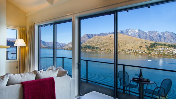 A modern hotel room with a large window overlooking a lake and mountains. There is a sofa with a red throw blanket, a table with a wine bottle, and a chair.