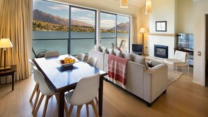 A modern living room with a large window overlooking a lake and mountains, a dining area with a table and chairs, and a cozy seating area with a fireplace.
