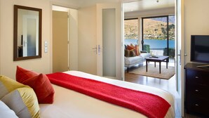 A bedroom with a large bed, a TV, and a balcony with a view of a lake and hills.
