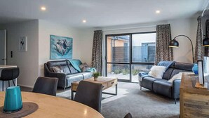 A modern living room with a sofa, coffee table, and dining table. There is a large window with curtains and a view of the outside. - Gallerie Coronet Summ (Queenstown)