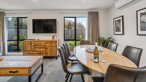 A dining area with a wooden table, leather chairs, a TV mounted on the wall, and large windows with curtains. - Gallerie Coronet Summ (Queenstown)