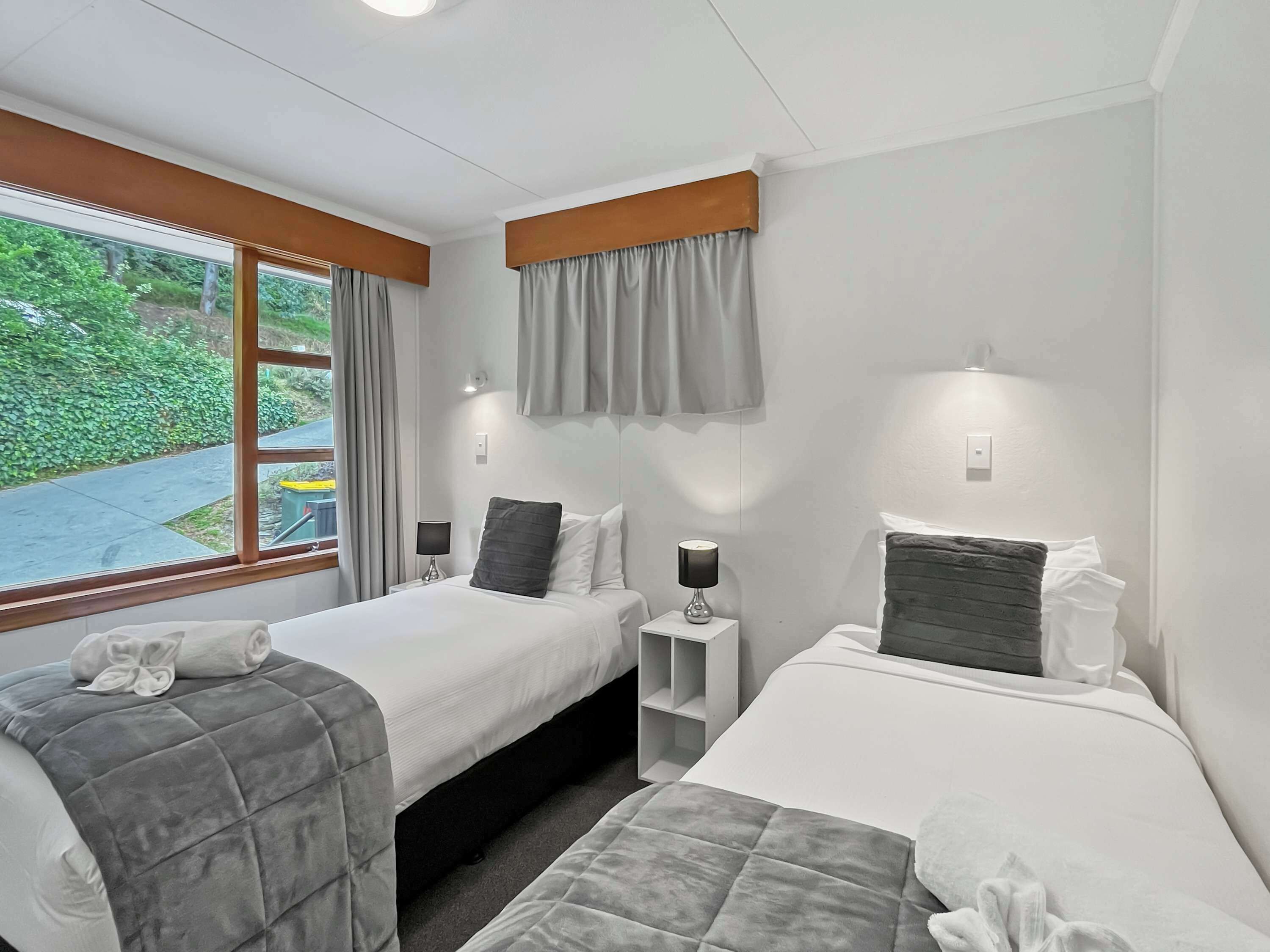 Ein Hotelzimmer mit zwei Betten, einem Vorhangfenster, Nachttischlampen und Blick auf Grünfläche.