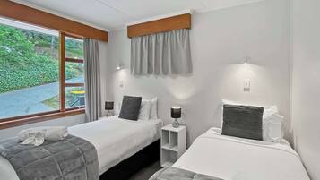 Ein Hotelzimmer mit zwei Betten, einem Vorhangfenster, Nachttischlampen und Blick auf Grünfläche.