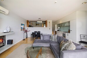 Een moderne woonkamer met een grijze bank, een glazen salontafel en een open haard. Er is een eethoek met een tafel en stoelen, en een keuken met witte kasten en een magnetron.