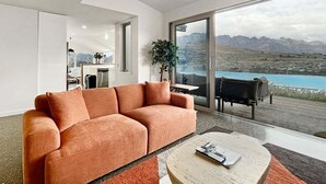 A modern living room with a sofa, a coffee table, and a view of mountains and a body of water through large glass doors. - Epic Views and Deck | Spacious Queenstown Home (Queenstown)