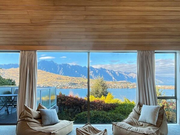 A room with a large window overlooking a lake and mountains, two beige sofas, and a wooden ceiling.