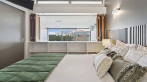 A modern bedroom with a large bed, a view of mountains through a window, and a built-in desk with a lamp. - Lakeview Premium 2 Bed Apartment Pounamu 203 (Queenstown)