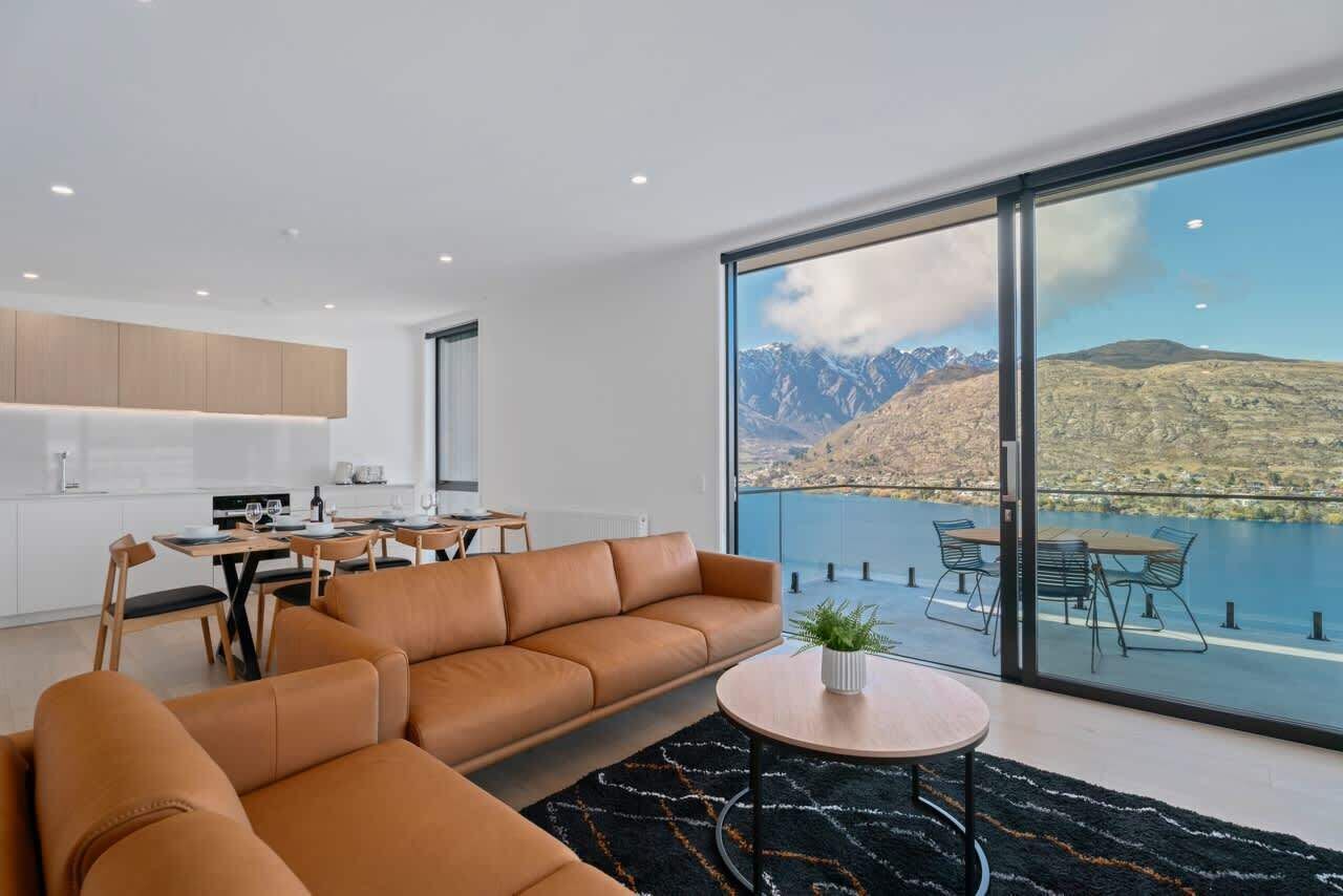 A modern living room with a large sofa, a dining table, and a view of a lake with mountains.