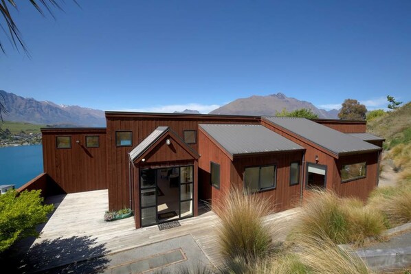 A modern wooden house with large windows and a deck overlooking a lake and mountains.
