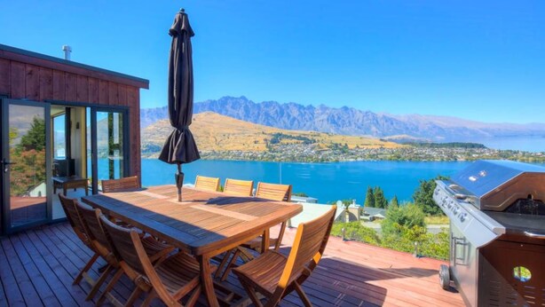 A wooden deck with a table and chairs, a grill, and a view of a lake and mountains.
