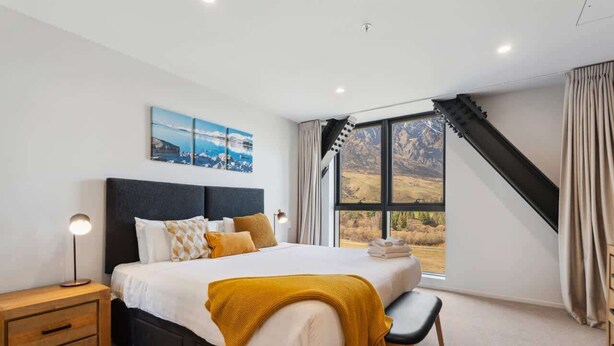A bedroom with a large bed, a nightstand, a bench, and a window with a view of a mountainous landscape. - Studio Getaway, Remarkables Views, Frankton (Queenstown)