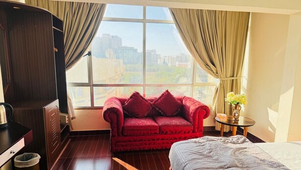 A hotel room with a large window, a red sofa, a bed, a small table with a vase, and a wardrobe.
