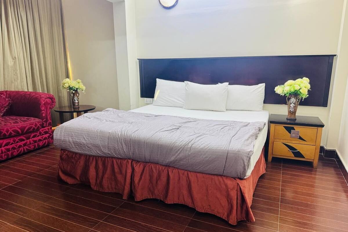 A hotel room with a large bed, a red armchair, a nightstand with a vase of flowers, and a wooden floor.